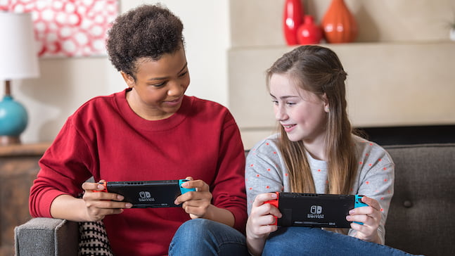 Deux femmes dans la même pièce connectent leurs consoles en mode sans fil local pour jouer.